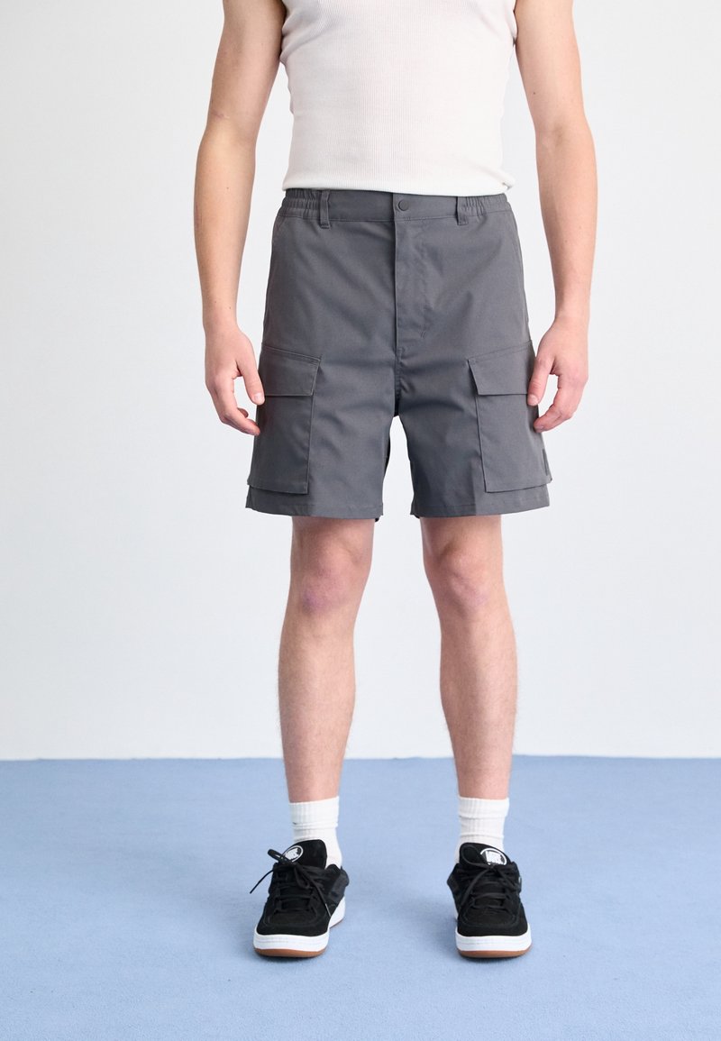 Personne debout portant un short cargo gris, des chaussettes blanches, des baskets noires et un débardeur blanc sur un fond uni.