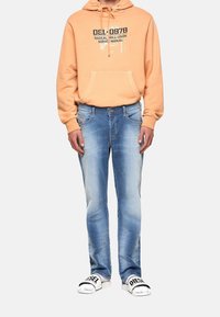 Sudadera naranja claro con un estampado gráfico, jeans de mezclilla azul claro con un acabado desteñido y sandalias blancas con la marca 'DIESEL'.