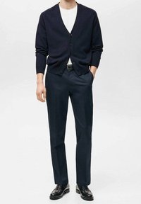 Tmavě modrý pletený cardigan s výstřihem do V a knoflíky, doplněný tmavými kalhotami a černými koženými botami, podtržený bílým tričkem.