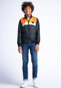 Zwarte en oranje lichte jas met witte streep, ritssluiting, elastische manchetten en zijzakken, gecombineerd met blauwe jeans en witte sneakers.