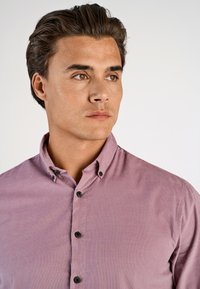 Camicia button-up di un leggero viola con colletto classico, caratterizzata da bottoni neri e un tessuto liscio e strutturato. Primo piano del design della camicia.
