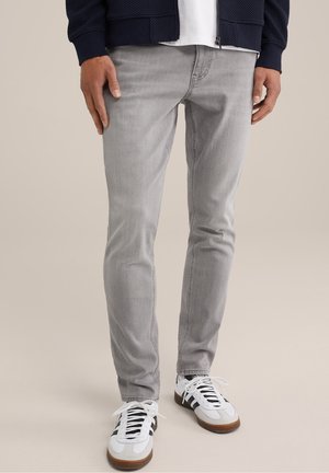 Mand iført lysegrå slim-fit jeans, hvide sneakers med sorte striber, en hvid skjorte og en navy textureret lynlåstrøje, står.