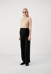 Beżowa prążkowana bluzka bez rękawów, czarne spodnie z wysokim stanem i szerokimi nogawkami z ozdobnym wiązaniem. Modelka nosi stylowe okulary przeciwsłoneczne i czarne buty.