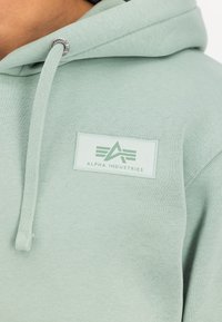 Világoszöld kapucnis felső, puha tapintással. Diszkrét logópajzs található a mellkasán, és fémes kiegészítők a húzózsinóron.