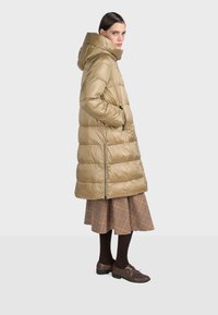 Pufferjacka i ljusbeige med huva, med horisontella quiltade sektioner och sidofickor, kombinerad med en rutig kjol och bruna skor.