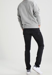 Persona vistiendo un sudadera gris, jeans negros ajustados y zapatillas Adidas blancas, de pie con las manos en los bolsillos contra un fondo liso.
