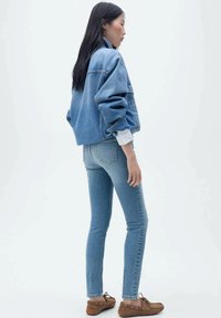 Modrá denimová oversized bunda s vysokým límcem, nošená přes bílou košili s dlouhým rukávem, kombinována s dobře padnoucími světle modrými džínami a hnědými botami.