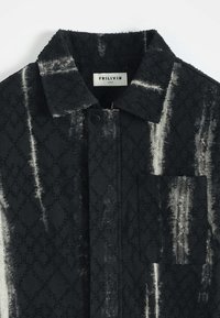 Veste à motifs noir et blanc en tissu texturé, avec un col, une fermeture à boutons sur le devant et une poche sur la poitrine gauche.