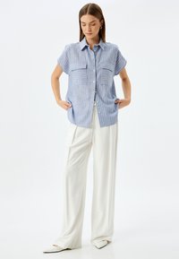 Camisa de rayas azul claro con mangas cortas y dos bolsillos frontales, combinada con pantalones anchos blancos y zapatos de punta.