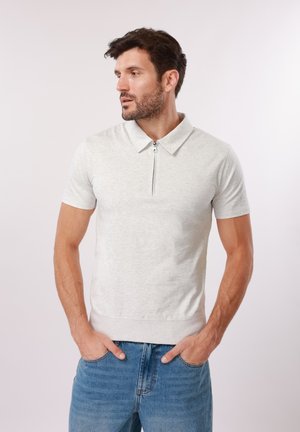 Man with dark hair and beard wearing light gray short-sleeve zip-up polo shirt and blue jeans, hands in pockets, looking left.