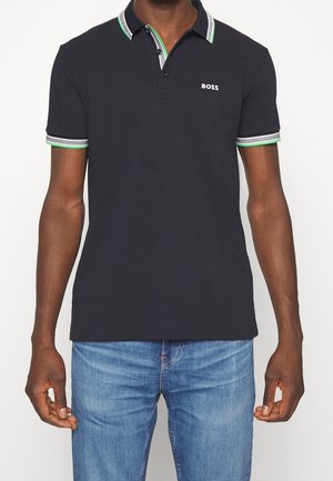 Polo negro con cuello y borde de las mangas a rayas verde, blanco y gris, logotipo "BOSS" en el pecho, combinado con jeans azules.