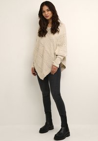Beige knitted poncho with a zigzag pattern, long sleeves, and a wide neckline, paired with black skinny jeans and chunky black boots.