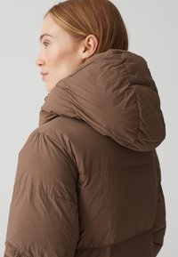 Braune Daunenjacke mit großer, plüschiger Kapuze. Texturiertes, leichtes Material mit gesteppten Abschnitten zur Isolierung. Einfaches Design, keine Muster.