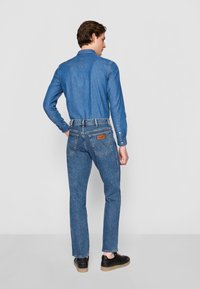 Chemise en denim pour homme de couleur bleu clair, à manches longues, avec des poignets à boutons et un design de gorge. Associée à un jean bleu slim avec des coutures contrastées et une patch logo.
