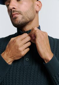 Donkergroen gebreide pullover met een ritssluiting bij de kraag, voorzien van een gestructureerd kabelpatroon en knoopsluiting. Handen die de rits aanpassen.