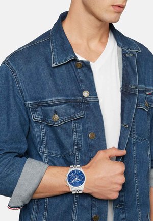 Sølvur med blå urskive og tre underdials, båret på et håndled. Modellen har en denimjakke med knaplukninger og en hvid skjorte under.