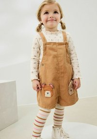 Salopette in corduroy color nocciola con toppe a forma di orso sulle tasche frontali, abbinate a una camicia lunga a maniche con stampa floreale crema e leggings a righe.