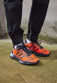 Sportovní boty s jasně oranžovým svrškem, černými detaily, texturovanou podrážkou a černými tkaničkami, spojené se zúženými černými tepláky.