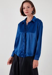 Satin blue blouse with a collared neckline, button front, and long sleeves; features a tied detail at the neckline and smooth texture.