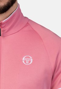 Rosa Zip-Jacke mit hohem Kragen, aus glattem Stoff gefertigt. Besitzt ein weißes Logodetail auf der linken Brust und kontrastierende Besätze.