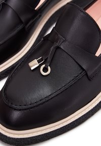 Mocassins en cuir noir avec détail de serrure et anneau argentés sur le dessus, bordure beige et semelles en caoutchouc noir texturé.