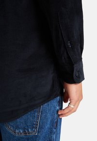 Navy corduroy shirt with vertical ribbing, buttoned cuffs, and a curved hem, paired with blue denim jeans featuring contrasting stitching.