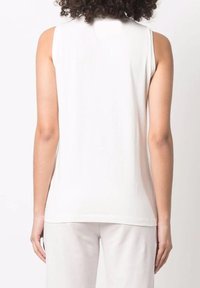 Persona con cabello rizado vestida con una blusa sin mangas blanca y pantalones blancos, vista de espaldas sobre un fondo blanco liso.