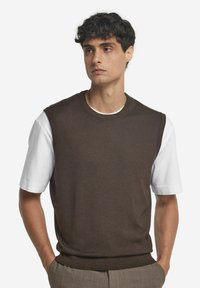 Brun stickad ärmlös tröja över en vit t-shirt, med rundhalsringning och ribbad nederkant, mot en enfärgad bakgrund.