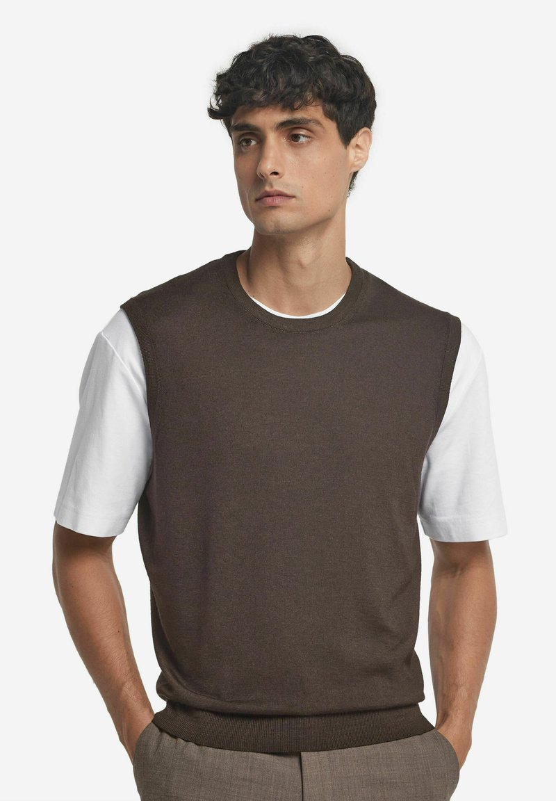 Brun stickad ärmlös tröja över en vit t-shirt, med rundhalsringning och ribbad nederkant, mot en enfärgad bakgrund.