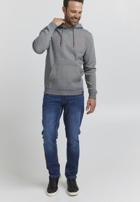 Hanorac gri cu dungi, dotat cu buzunar kangaroo, șnururi ajustabile și detaliu cu logo, asortat cu blugi albaștri și adidași negri.