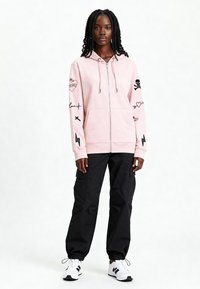 Felpa con cappuccio rosa con zip e ricami sulle maniche, abbinata a pantaloni cargo neri e sneakers bianche. Tessuto morbido e vestibilità comoda.