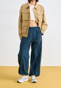 Beżowa krótka kurtka z wieloma kieszeniami, zestawiona z luźnymi spodniami w ciemnym odcieniu teal z elastycznym pasem i logo Nike. Srebrne sneakersy.