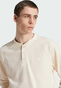 Camisa de manga larga color crema con un tejido de punto tipo waffle, cuello redondo, tres botones y un logo blanco bordado.