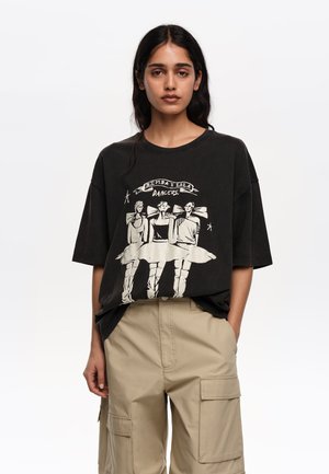 Kobieta w czarnej, oversize koszulce z ilustracją trzech baletnic i beżowych spodniach typu cargo, stojąca na jednolitym białym tle.