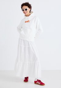 Vit hoodie med orange Levi's-logga, vit skandinavisk maxiklänning i flera lager och röda sneakers med vita ränder. Solglasögon och lockigt hår fulländar looken.