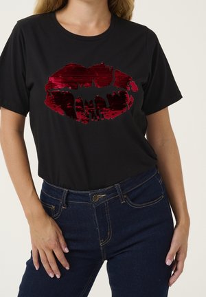 Vrouw die een zwart T-shirt draagt met een glanzend rood lippenontwerp en donkerblauwe jeans, poseert met één hand op de heup en één langs het lichaam.