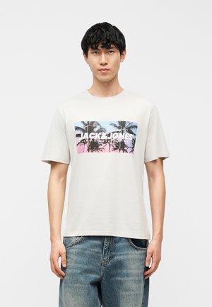 Jonge man met een wit T-shirt met palmboomprint en tekst "JACK & JONES", gecombineerd met blauwe spijkerbroek, staand tegen een effen achtergrond.