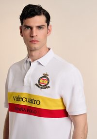 Camisa polo blanca con rayas horizontales amarillas y rojas. Escudo bordado con una corona, el logo "valecuatro" y el número "2" en la manga.