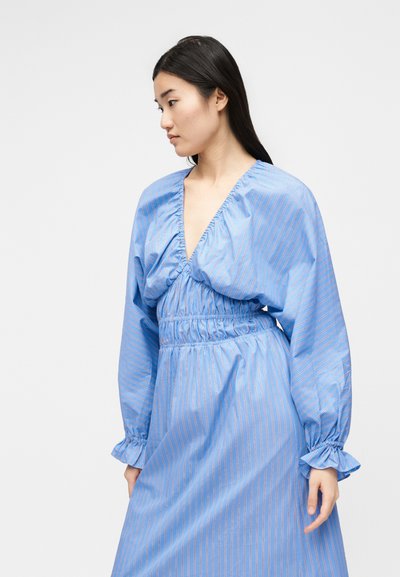 Νεαρή γυναίκα με μακριά σκούρα μαλλιά, που φοράει μπλε ριγωτό φόρεμα με βαθύ V-λαιμόκοψη, σούρωμα στη μέση και βολάν στα μανίκια, κοιτάζει προς τα αριστερά.