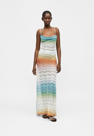 Γυναίκα που φοράει αμάνικο, μακρύ φόρεμα με διαβαθμίσεις πολλαπλών χρωμάτων και σχέδιο κυμάτων, μαζί με σανδάλια με λουράκια, στέκεται μπροστά σε απλό φόντο.