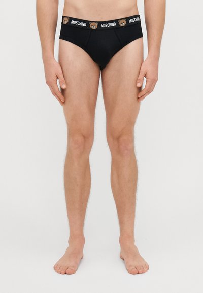 Črne bombažne hlačke s kontrastnim pasom, ki vsebuje logotip medveda in besedilo "MOSCHINO." Klasičen kroj hlačk, gladka tekstura.