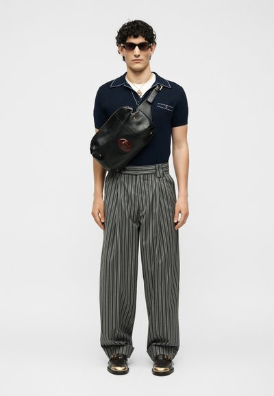 Άνδρας που φοράει μπλε μπλούζα με κοντό μανίκι, ριγέ φαρδιά παντελόνια, σκουρόχρωμα γυαλιά ηλίου, μαύρη τσάντα χιαστί και μαύρα παπούτσια με χρυσά μύτα, στέκεται μπροστά σε λευκό φόντο.