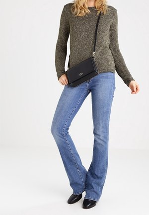 Nainen, joka pukeutunut harmaaseen pitkähihamaiseen neulepaitaan, sinisiin bootcut-farkkuihin, mustiin teräväkärkisiin kenkiin ja mustaan olkalaukkuun, seisoo valkoista taustaa vasten.