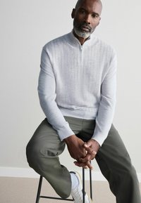 Hellgrau gestrickter Pullover mit Reißverschlusskragen, strukturiertem Muster und gerippten Bündchen, kombiniert mit olivgrüner Hose und weißen Sneakers.