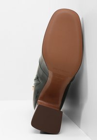 Stivale da donna alla caviglia in pelle verde scuro, mostrato dal suola con un tacco quadrato marrone e il nome del marchio impresso.