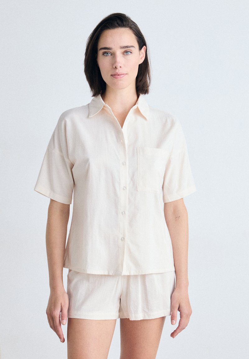 Camisa de manga corta de color beige claro con botones en la parte delantera y un bolsillo, combinada con pantalones cortos a juego, confeccionada en una tela suave y lisa.