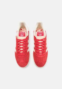Röda mockaskor med vita ränder, snörning och vadderad krage. Har röd logotyp på tungan och mönstrad innersula. Gummisula.