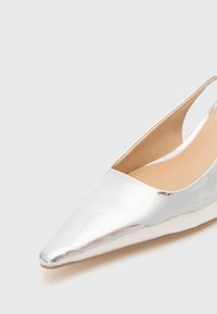 Chaussure argentée métallique à bout pointu avec un design épuré, côtés ouverts et texture lisse. Le talon est fin et allongé pour un look moderne.