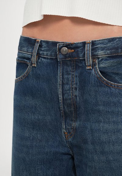 Kék farmernadrág derékrésze gombbal és narancssárga varrással, fehér, rövidített bordázott felsővel viselve, egyszínű háttér előtt.