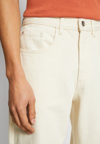 Beige cotton pants with a classic five-pocket design, featuring a metal button closure and minimal stitching detail.